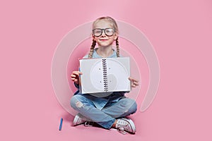 Happy little girl in glasses showing exam paper on pink background