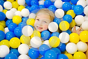 Happy little girl having fun in ball pit in kids indoor play center. Child playing with colorful balls in playground ball pool.