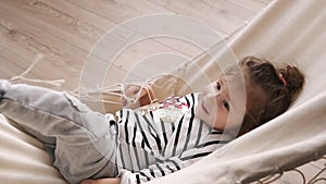 Happy little girl having fun indoors, lying in hammock, and waving her legs. Smiling child