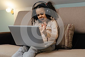 The happy little girl in headphones sitting at the sofa with a laptop