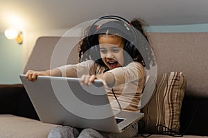 The happy little girl in headphones sitting at the sofa with a laptop