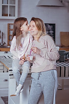 Happy little girl and her beautiful young mother have breakfast together in a white kitchen. Daughter kisses her mom. Maternal