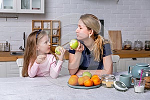 Happy little girl and her beautiful young mother have breakfast together in a white kitchen. They are having fun and eating apples