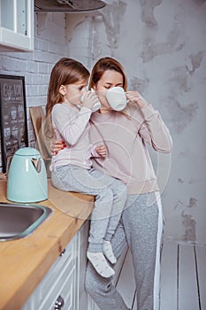 Happy little girl and her beautiful young mother have breakfast together in a white kitchen. They hug and drink tea. Maternal care