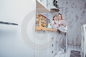 Happy little girl and her beautiful young mother have breakfast together in a white kitchen. They hug and drink tea. Maternal care