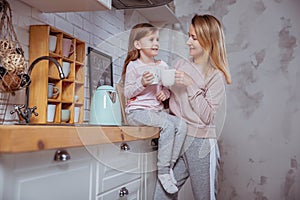 Happy little girl and her beautiful young mother have breakfast together in a white kitchen. They hug and drink tea. Maternal care