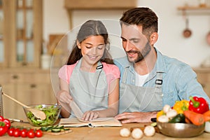 Happy little girl and her father reading coolinary book, choosing recipe or checking ingredients, preparing together