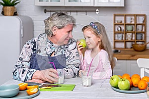 Happy little girl and her grandmother have breakfast together in a white kitchen. They are having fun and playing with fruits.