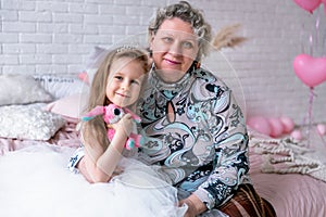 Happy little girl and her grandmother are sitting together and hugging in the bedroom. They are smilling and kissing. Maternal