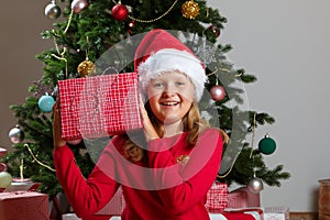Happy little girl holding a box with a gift. Child in a Santa hat against the background of a Christmas tree