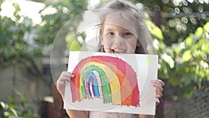 Happy little girl holding a children's rainbow drawing outdoor. Summer sunny day