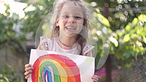 Happy little girl holding a children's rainbow drawing. Summer sunny day