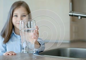 Happy little girl holding a glass of water. Selective focus on child hand with glass of water in foreground. Hydration. Water