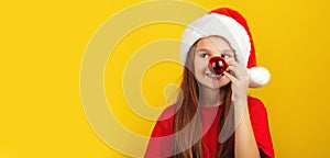 Happy little girl holding red christmas tree ball in front of his nose, wearing Santa hat posing against yellow studio wall
