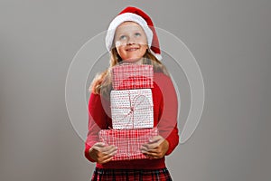 Happy little girl holding a stack of Christmas gifts isolated gray background. A child in a Santa hat looks up dreamily