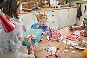Happy little girl with an inflatable crown celebrating her birthday, sitting by the table with guests, recieving gifts from her