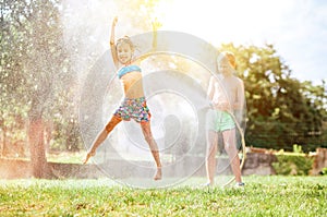 Happy little girl jumps under water, when brother pours her from garden hose. Hot summer days activity