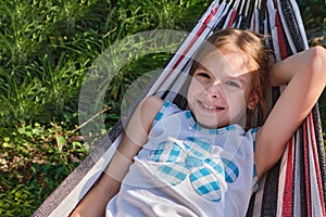 A happy little girl lies in a hammock in the garden in the summer. A child in a hammock during the holidays smiles in