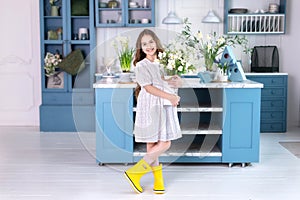 Happy little girl with long hair in a white dress and in yellow rubber boots with a bouquet of flowers, stands near kitchen