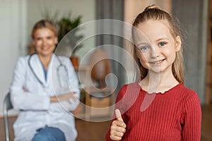 Happy little girl at pediatrician clinic with female doctor on background smiling at camera and showing thumb up