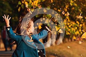 Happy little girl playing with autumn leaves. Cute child having fun in park. Stylish baby girl in blue coat throws autumn leaves o