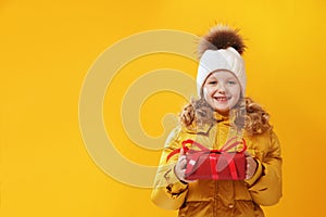 Happy little girl preschooler in a winter jacket and in a knitted hat is holding a box with a gift.