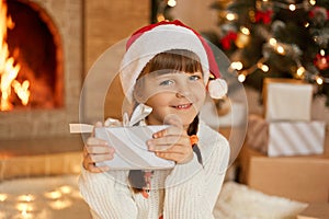 Happy little girl with present at Christmas Eve, sitting near xmas tree and fireplace, charming smiling child looking at camera