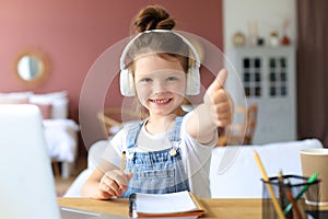 Happy little girl pupil study online using laptop at home, smiling small child show thumb up recommend class or lesson