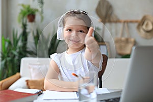 Happy little girl pupil study online using laptop at home, smiling small child show thumb up recommend class or lesson