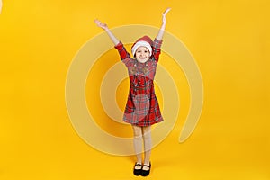 Happy little girl raised her hands up. Child in santa claus hat and red dress on a yellow background