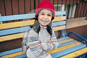 A happy little girl in a red winter hat is sitting on a colorful bench outside and waiting for her friend. Pretty kid takes a rest