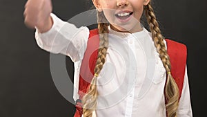 Happy little girl with rucksack showing thumbs up against blackboard, studying