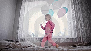 Happy little girl is running near the window on the bed in the nursery with the bunch of air balloons in her hands and