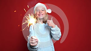 Happy little girl in Santa Claus cap holding bengal light on the red background