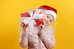 Happy little girl in santa hat is holding christmas gift. Baby in a pink jacket on a yellow background