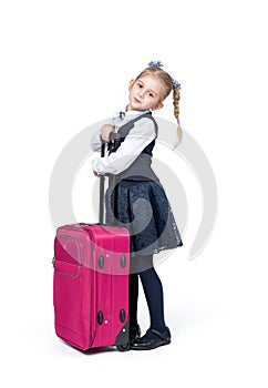 A happy little girl in a school uniform stands with a large pink suitcase, isolated on a white background