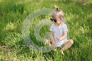 Happy little girl sits on the grass outdoors. Cute baby girl in white bodysuit and sunglasses on the backyard. Adorable