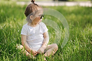 Happy little girl sits on the grass outdoors. Cute baby girl in white bodysuit and sunglasses on the backyard. Adorable