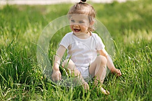 Happy little girl sits on the grass outdoors. Cute baby girl in white bodysuit and sunglasses on the backyard. Adorable