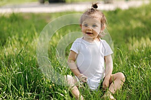 Happy little girl sits on the grass outdoors. Cute baby girl in white bodysuit and sunglasses on the backyard. Adorable