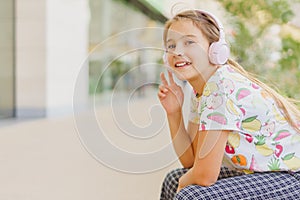 Happy little girl sits in town on a bench and listens to music through wireless headphones
