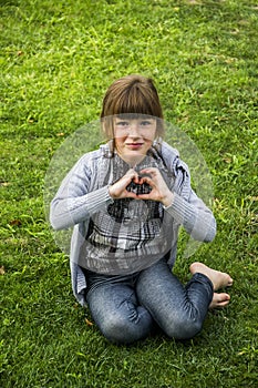 Happy little girl sitting on the grass and make handly sign of heart