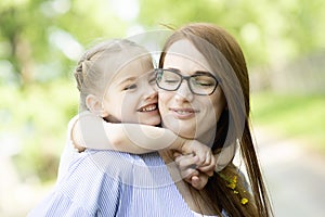 Happy little girl sitting on her mom shoulders or back on a sunny summer day and kissing her mother cheek