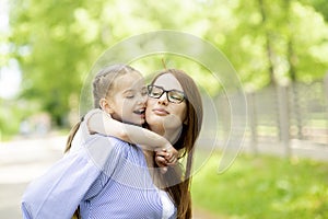 Happy little girl sitting on her mom shoulders or back on a sunny summer day and kissing her mother cheek