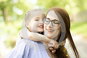 Happy little girl sitting on her mom shoulders or back on a sunny summer day and kissing her mother cheek