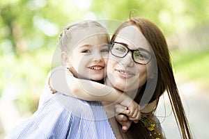 Happy little girl sitting on her mom shoulders or back on a sunny summer day and kissing her mother cheek