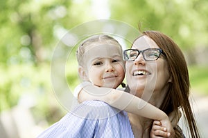 Happy little girl sitting on her mom shoulders or back on a sunny summer day and kissing her mother cheek