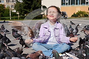 Happy little girl sitting in lotus pose with her eyes closed, feeds flock of flying pigeons and birds in park square