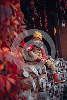 Happy little girl sitting at pile of wood with flower, surrouded by red leaves, during beautiful autumn day.