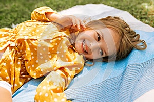 Happy little girl smiling broadly and lying on the blanket at the grass, enjoying summer day outdoors. Adorable child having fun